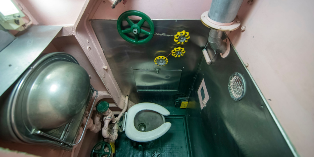 Ubåds kompakte toiletfaciliteter, der viser et metaltoilet, forskellige rør, en ventil og advarselsmærker, submarine