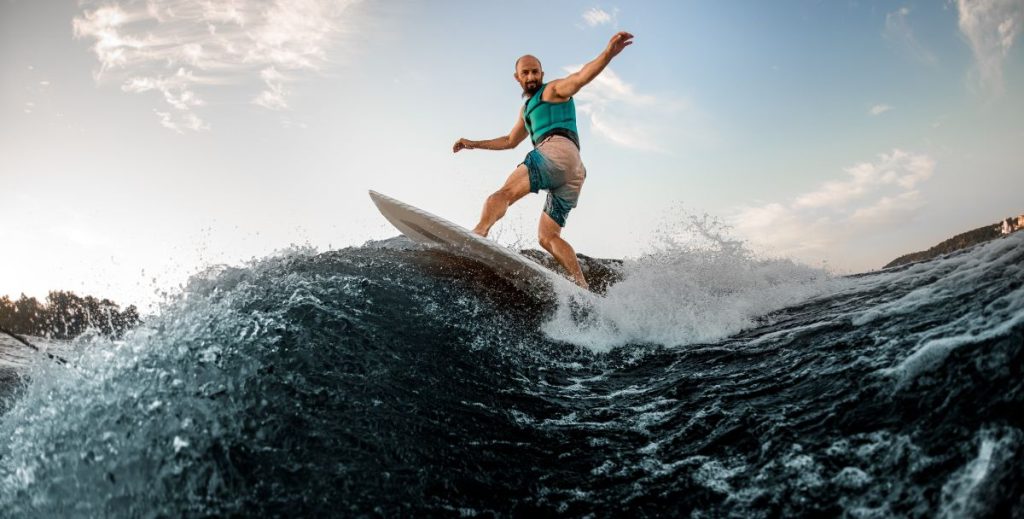 sportive man on turquoise lifejacket is surfing on surfboard trails behind a boat.