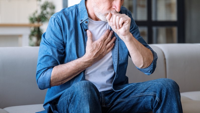 Senior man sitting on sofa at home and holding hand on chest. Male having asthma attack, difficulties with breathe, feeling severe pain or dyspnea