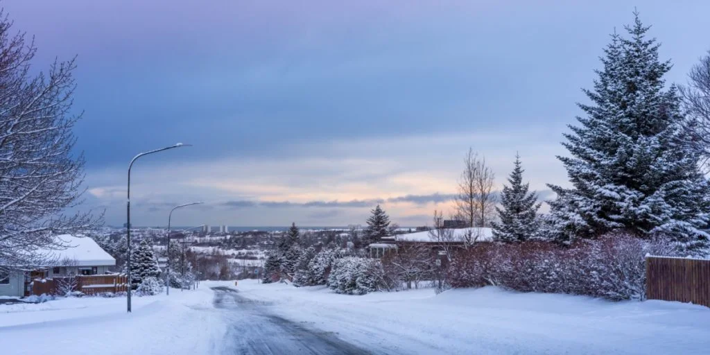 Empty snowy street in Reykjavik suburbs on a dark winter day. Residential buildings, cars driving down the road. Gray, stormy clouds in the sky.