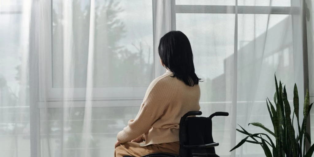 A woman sits in a wheelchair, facing a window, looking out at the view.