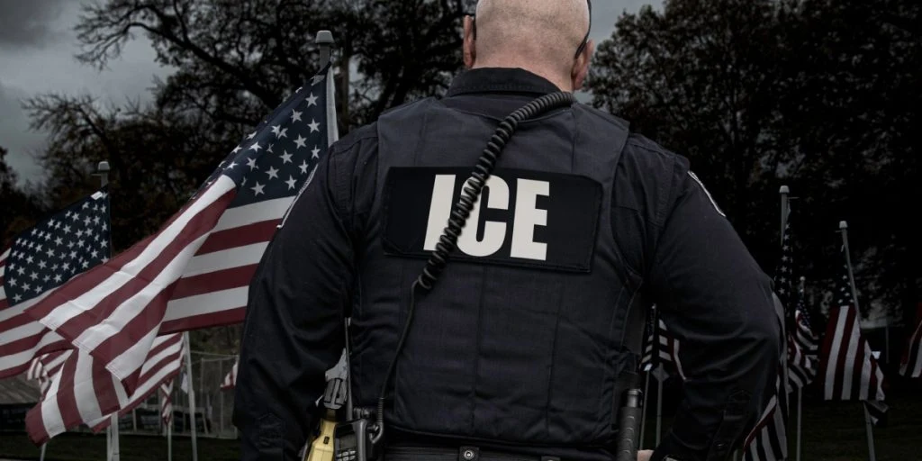 An immigration (ICE) officer stands in front of rows of US flags