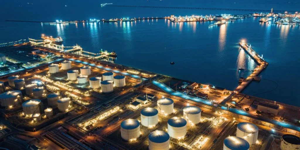 Aerial view of industrial oil terminal port at dusk with illuminated refinery tanks, cargo holds and shipping infrastructure reflecting on calm sea, capturing global energy logistics and maritime industry