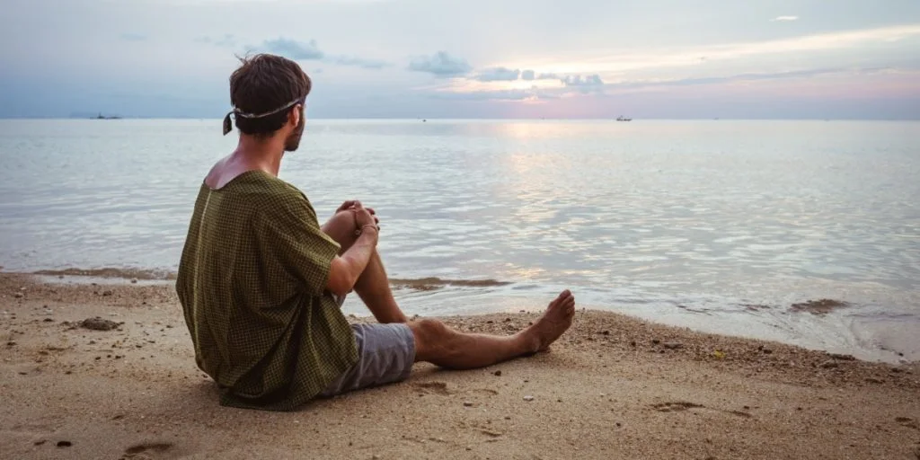 Young man meditating on the beach sitting on the sand looking at the sea