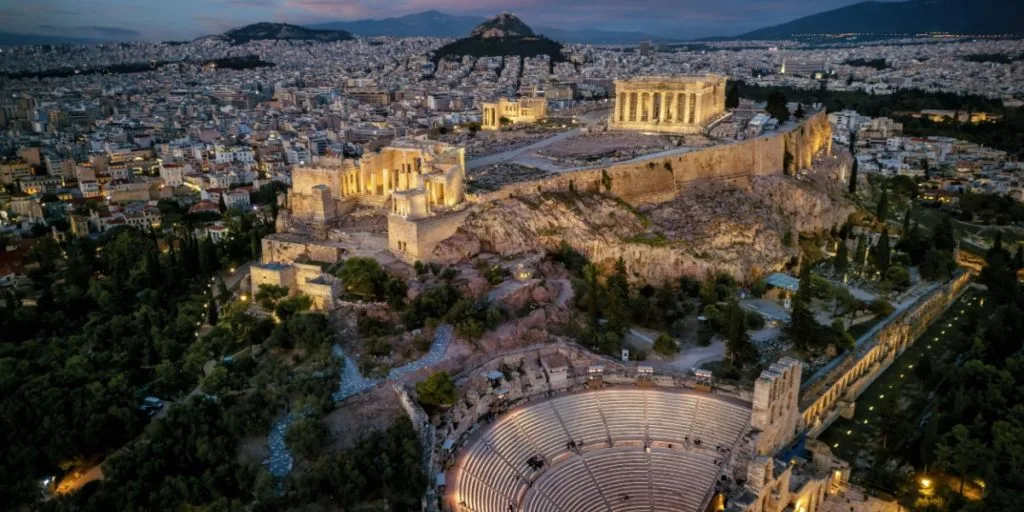 Aerial view of illuminated, ancient ruins at the Acropolis of Athens, Greece, and Odeon of Herode theatre during dusk
