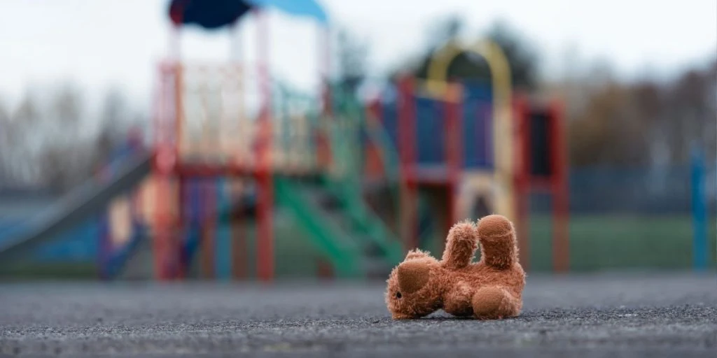 Abandoned teddy bear lies on empty playground with blurred colorful equipment in the background.