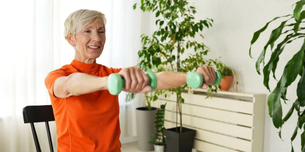 An elderly woman sits on a chair and lifts green dumbbells while smiling. She works out in a bright living room with plants, promoting strength, fitness and an active lifestyle.