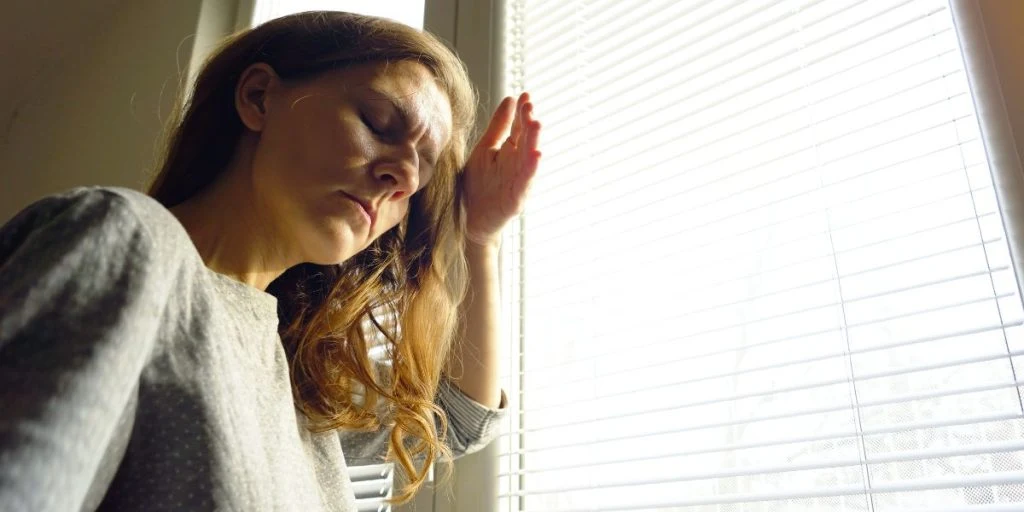 A woman stands near the blinds, closing her eyes and shielding her face from the sunlight with her hand. Highlighting photophobia, overstimulation, acute migraine symptoms and neurological tension.