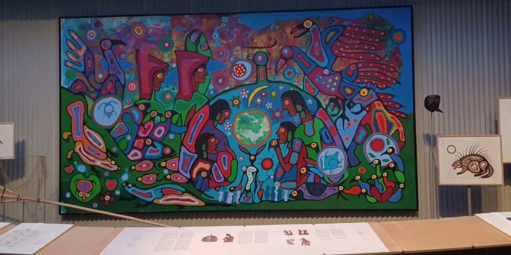 A large, colorful painting featuring First Nations imagery in a graphic style is displayed on a museum wall.
