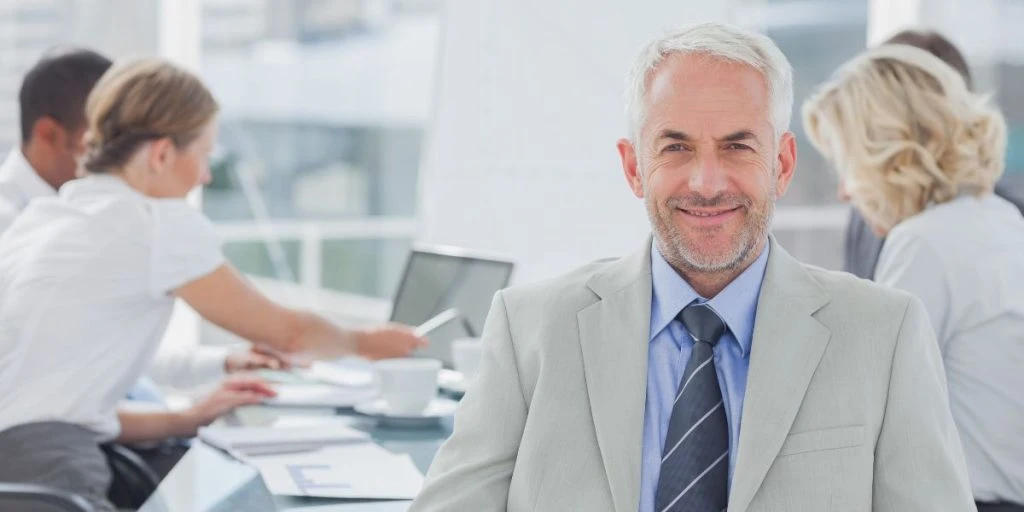 Charismatic businessman posing in the boardroom while colleagues are working behind