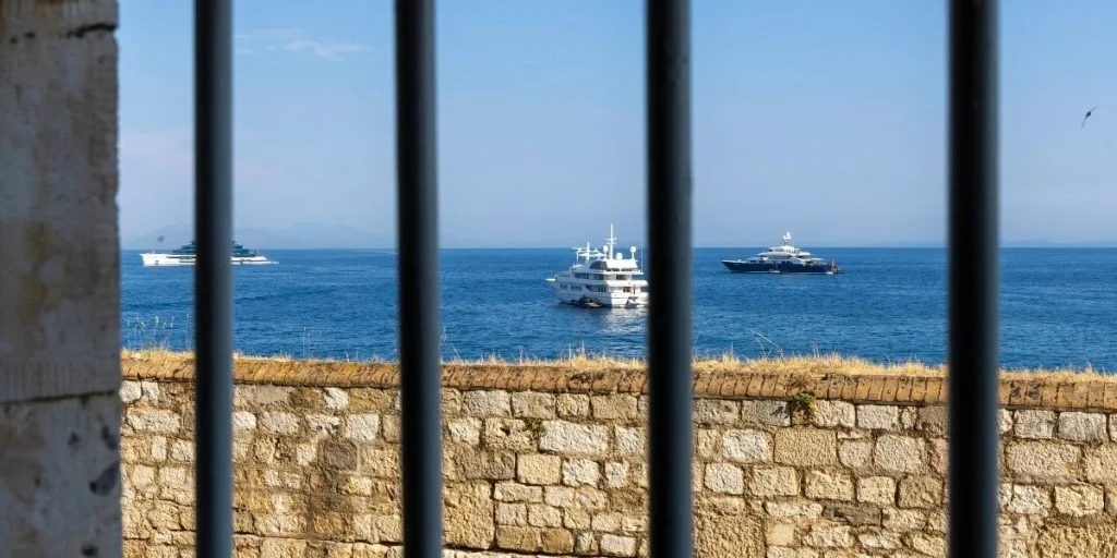 Corfu, Greece - Old stone wall and window bars frame luxury yachts on the calm Ionian Sea