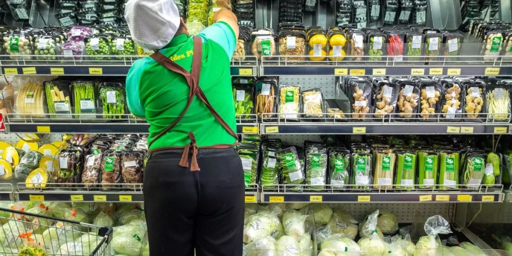 A supermarket employee stocks produce in a grocery store. The image is illustrative.