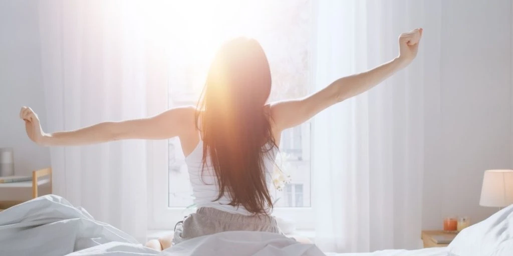 Brunette wakes up in the morning, stretches in bed, sun shines on her from the big window. Happy young girl greets new day with warm sunlight Flare