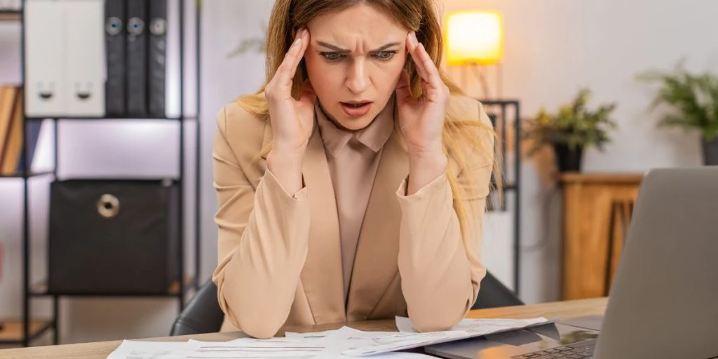 Middle-aged businesswoman at home office looking at bills invoices receipt on laptop seeing costs going up frowning confused and unhappy